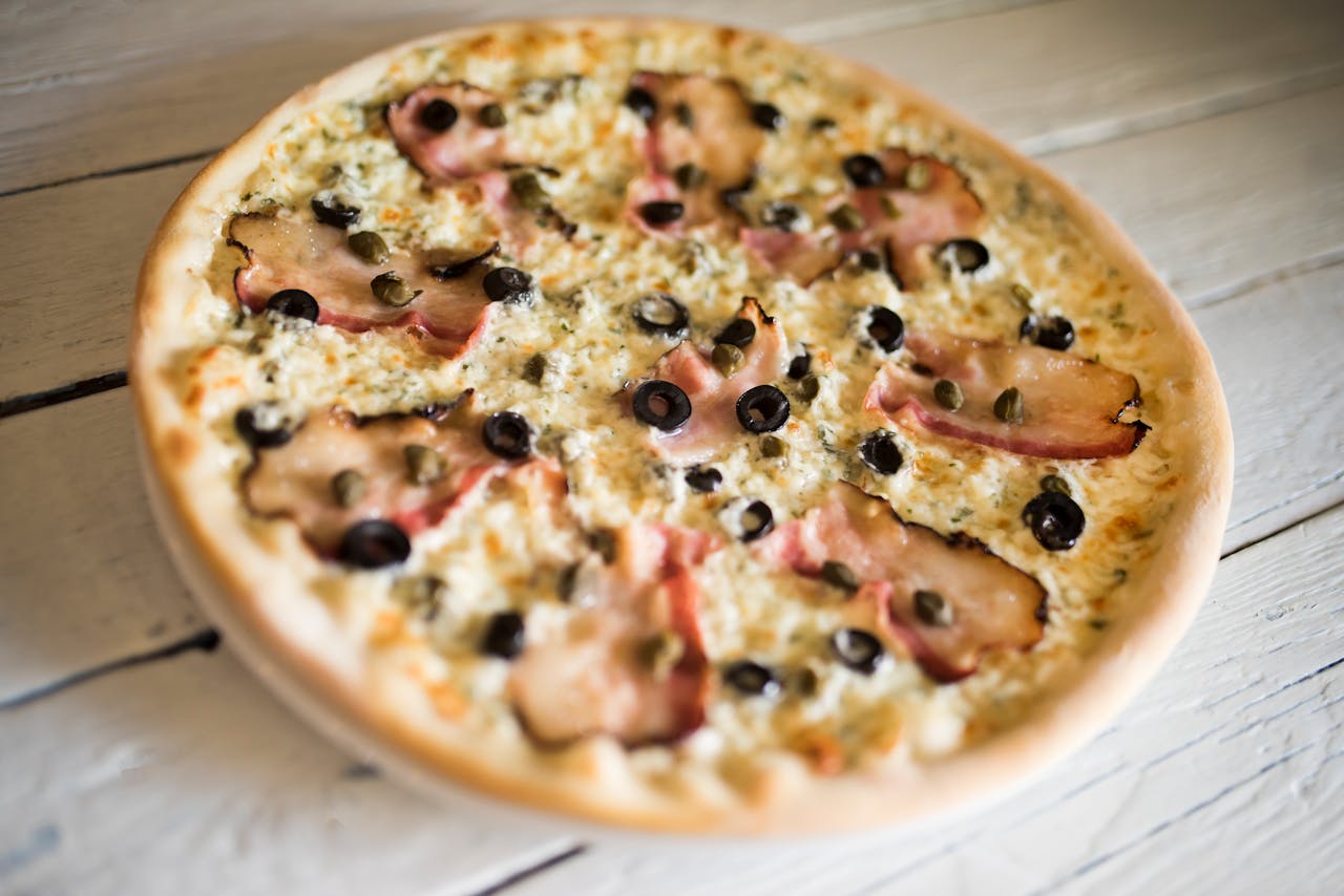 home-hero-1 A mouth-watering white cheese pizza topped with olives, capers, and ham on a rustic wooden table.